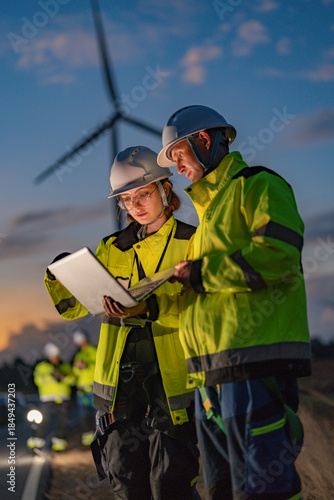 Male and female engineers in safety gear inspecting wind turbines at dusk. Female engineer holding a walkie-talkie while her colleague uses a laptop for green energy monitoring.