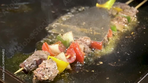 A close-up of a meat and vegetable kebab cooking on a pan, oily griddle. The skewer holds chunks of seasoned meat