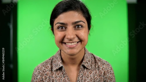 A young woman with dark hair smiles broadly in a studio setting with a green screen background and professional lighting equipment visible