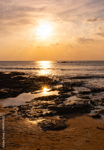 Dramatic sunset with sun reflection on a rocky beach in Khao Lak Thailand. Golden light illuminates the rocks, sea and sky, capturing a tranquil tropical coastal landscape at dusk.