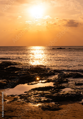 Dramatic sunset with sun reflection on a rocky beach in Khao Lak Thailand. Golden light illuminates the rocks, sea and sky, capturing a tranquil tropical coastal landscape at dusk.