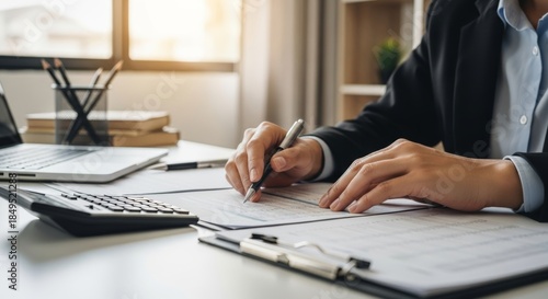 Businesswoman in formal attire reviewing documents at a desk with a laptop and calculator, office setting