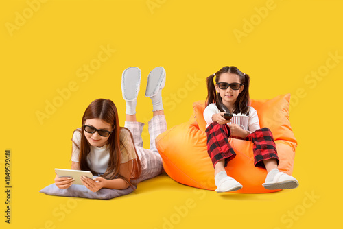 Cute Asian sisters in 3D glasses watching cartoons on yellow background