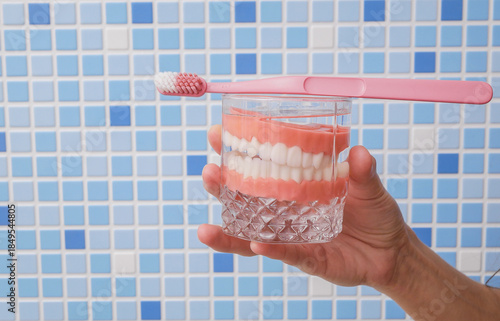 Hand holding dentures in glass and toothbrush against blue bathroom tiles