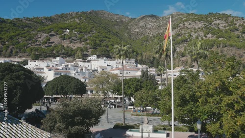 Street view in Mijas Pueblo, Spain, showing a green mountain, traditional white buildings, and a Spanish flag fluttering on a sunny day.