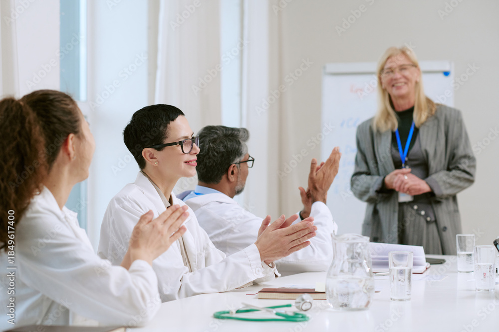 Fototapeta premium Middle aged Caucasian woman standing and smiling while presenting to diverse group of young adult and middle aged doctors, sitting at table and applauding during medical seminar