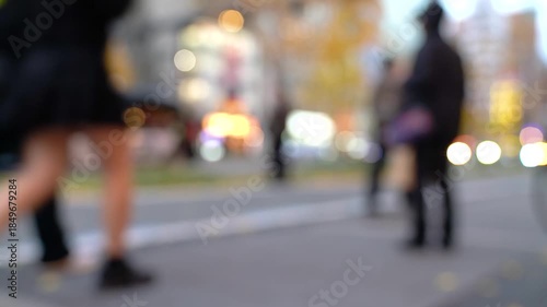 Blurred street scene with people walking and car lights creating bokeh effect. Atmospheric urban background, motion blur conveys movement and anonymity. Abstract city life concept.