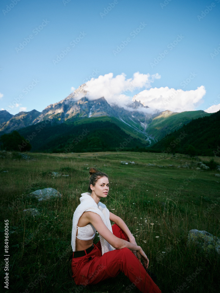 Fototapeta premium Mountain landscape with blue sky and clouds, a woman sits in a meadow wearing red pants and a white top, outdoor fashion in scenic nature.