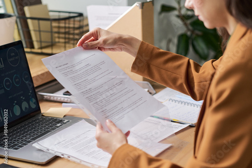 Young adult Caucasian woman reviewing printed documents at desk, holding paper in hand while working on business project with open laptop and financial charts visible on screen