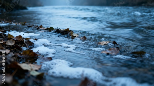 A tranquil riverside scene featuring gently flowing water along the shore, with fallen autumn leaves and soft foam resting at the edge.
