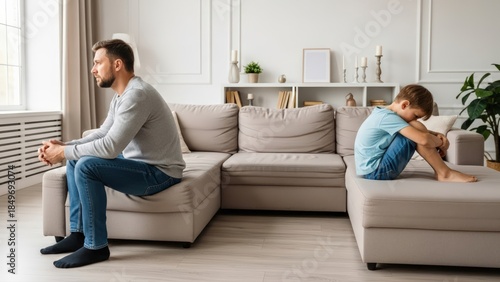 Unhappy father and young son sit apart on a modern beige sofa, silent tension highlighting a generation gap after a family quarrel