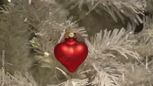 A red heart-shaped ornament hangs on a white Christmas tree. The ornament features a gold cap and is surrounded by snowy branches.