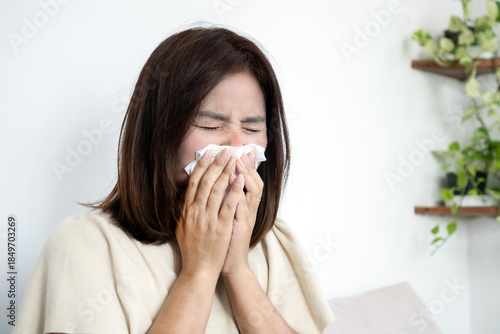 Photos Asian woman sneezing into a tissue, suffering from seasonal allergies, common cold, or flu symptoms at home