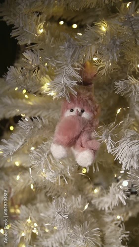 A cute pink plush ornament hangs on a white Christmas tree adorned with warm lights. The tree has a snowy appearance, creating a festive atmosphere.
