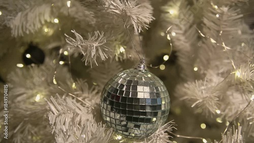 A close-up of a shiny disco ball ornament hanging on a white Christmas tree, surrounded by warm glowing lights.