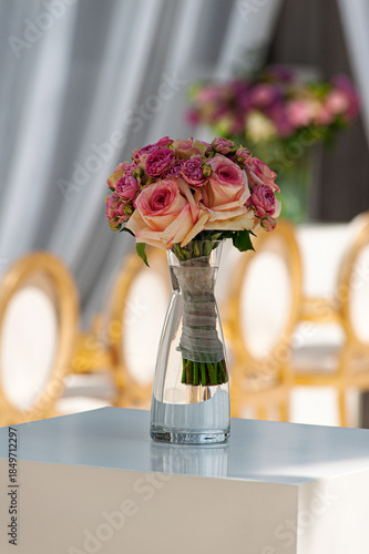 A romantic pink rose bouquet arranged as a centerpiece in a clear glass vase, wrapped with twine. Soft, elegant decor creates a refined wedding or special-event ambiance around a stylish table setting
