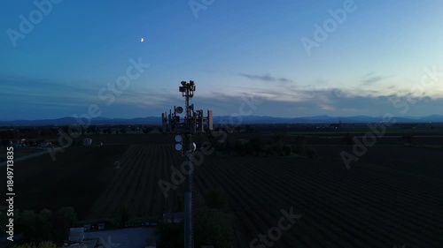 Aerial footage of a tall telecommunication tower equipped with antennas and satellite dishes, transmitting signals against a clear dark blue sky at dusk in a rural landscape