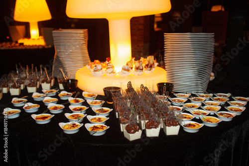 A stylish dessert display at a dimly lit event featuring cream cups, chocolate treats, and bite-sized pastries arranged on a black tablecloth, with warm yellow lighting and stacked white plates adding