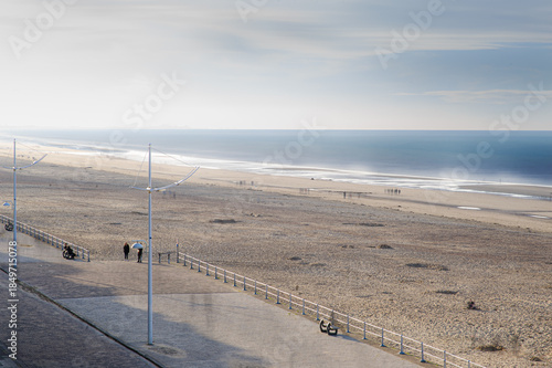 A sunlit coastal scene featuring a wide promenade along the beach. Calm waves, blue ocean, and distant walkers create a peaceful seaside atmosphere.
