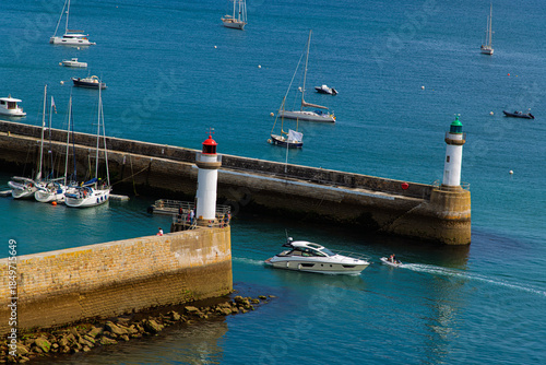 A tranquil harbor scene with boats docked near stone breakwaters, two lighthouses guarding the blue sea. A motorboat glides past as sailboats anchor, creating a bright, nautical travel vibe.