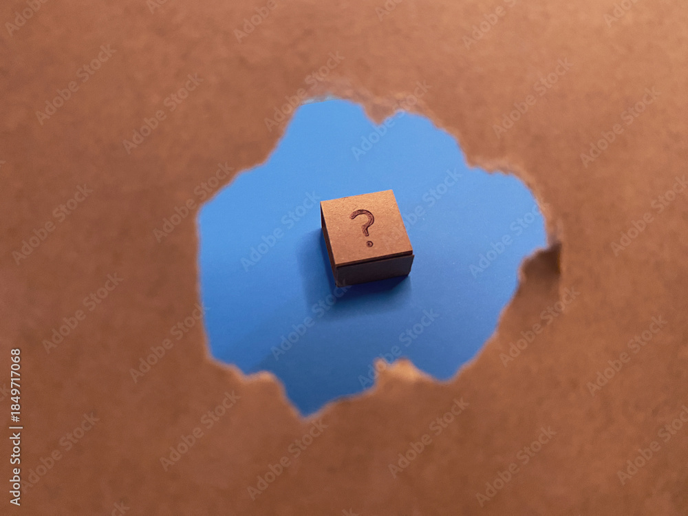 Fototapeta premium Conceptual image featuring a question mark on a small block, revealed through a hole in brown paper against a blue background. Ideal for FAQs.
