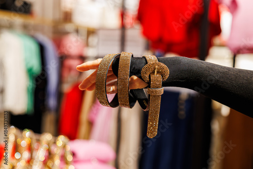 Wallpaper Mural A glittery gold rhinestone-studded belt with a matching round buckle is elegantly draped over a hand in a clothing store with colorful garments in the background Torontodigital.ca