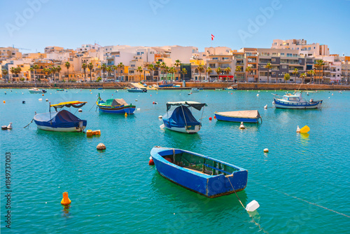 Marsaxlokk Harbour, Malta, is busy with colorful luzzu fishing boats in turquoise waters. Quaint waterfront buildings complete sunny Mediterranean scene