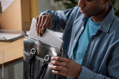 Fotografi Young adult Black man placing laptop into leather briefcase, sitting at desk in
