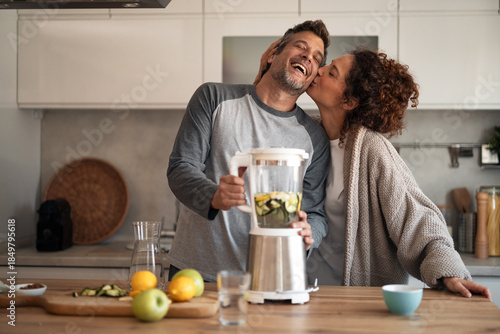 Joyful Couple In A Modern Kitchen Blending Fruit, Sharing A Kiss And Laughing Together
