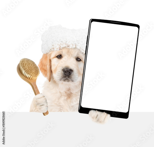 Golden retriever puppy with soap foam on it head holds shower brush and big smartphone with white blank screen in it paw above blank white banner. isolated on white background