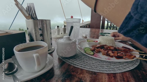 Low angle view taking cutlery to get ready to eat pork barbecue skewers served with rice and coffee in a restaurant with the view of the foggy landscape, candid authentic moment