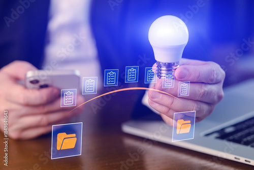 Business person holds light bulb while using smartphone and laptop with document icons above