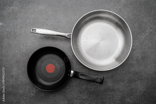 Top view of professional stainless steel and black non-stick frying pans on a dark grey stone background. Kitchenware and cookware concept