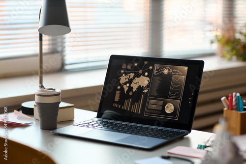 Wallpaper Mural Open laptop displaying data analytics dashboard with world map and charts on screen, sitting on office desk near reusable coffee cup, lamp, and stationery, blinds covering window Torontodigital.ca