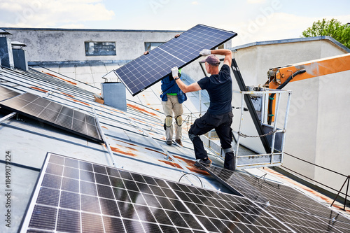 Workers building solar panel system on metal rooftop of house with assistance of crane lift. Two men installers carrying photovoltaic solar module outdoors. Renewable energy generation concept.