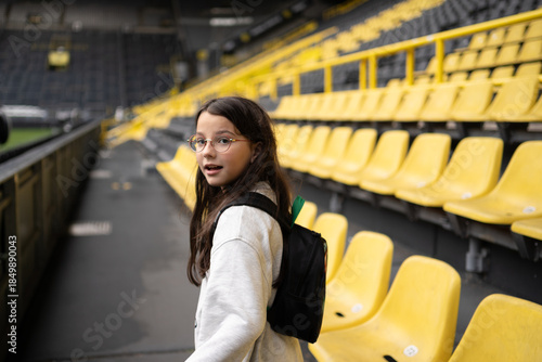 Smiling teenage girl in glasses with backpack on empty football stadium, education travel and leisure concept