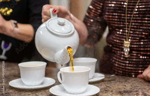 Woman pouring tea from a classic white ceramic teapot into teacups.