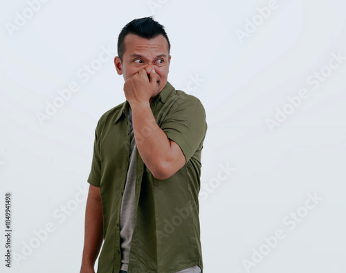 Asian man isolated over white background smelling something stinky and disgusting, intolerable smell, holding breath with fingers on nose. Bad smells concept.