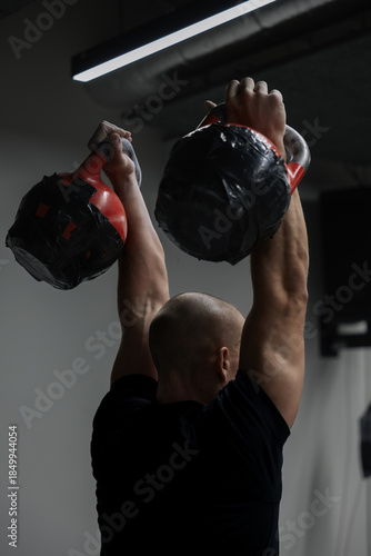 Rear view of strong male athlete performing double kettlebell jerk during strength training in gym. Power, endurance, functional fitness and professional sport concept.