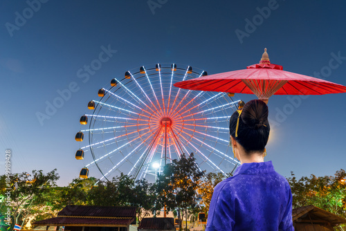 A female tourist in traditional Thai dress holds an umbrella while visiting a Ferris wheel in Nakhon Phanom Province, Thailand.