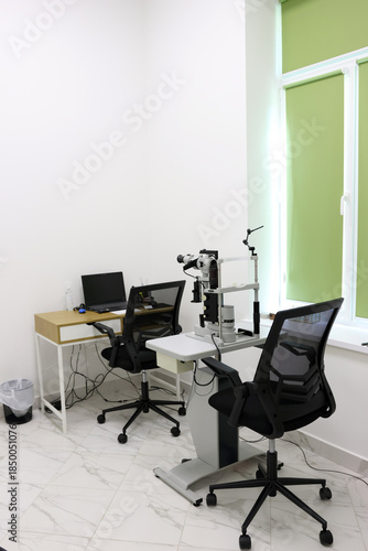 Interior of an ophthalmologist's office with a slit lamp biomicroscope, a table with a laptop in a bright and clean medical examination room.