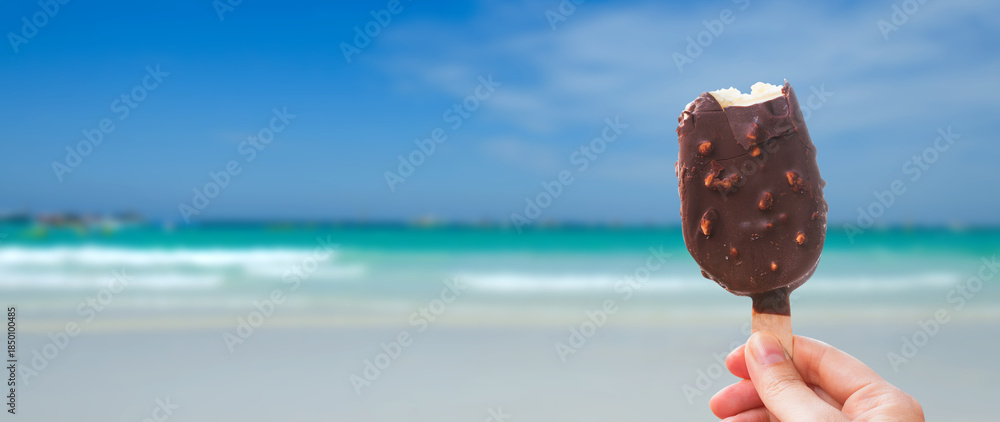 Obraz premium Chocolate covered ice cream bar in the summer at the beach, during the holidays. Hand holds delicious chocolate and nuts almond ice cream bar on a stick.