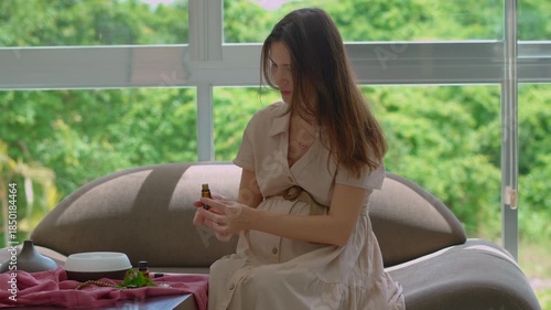 Pregnant woman over 40 sitting on a couch by a sunlit window enjoying scent and relaxation from a working electric aroma diffuser. Wellness, pregnancy care and relaxation lifestyle concept. Slowmotion