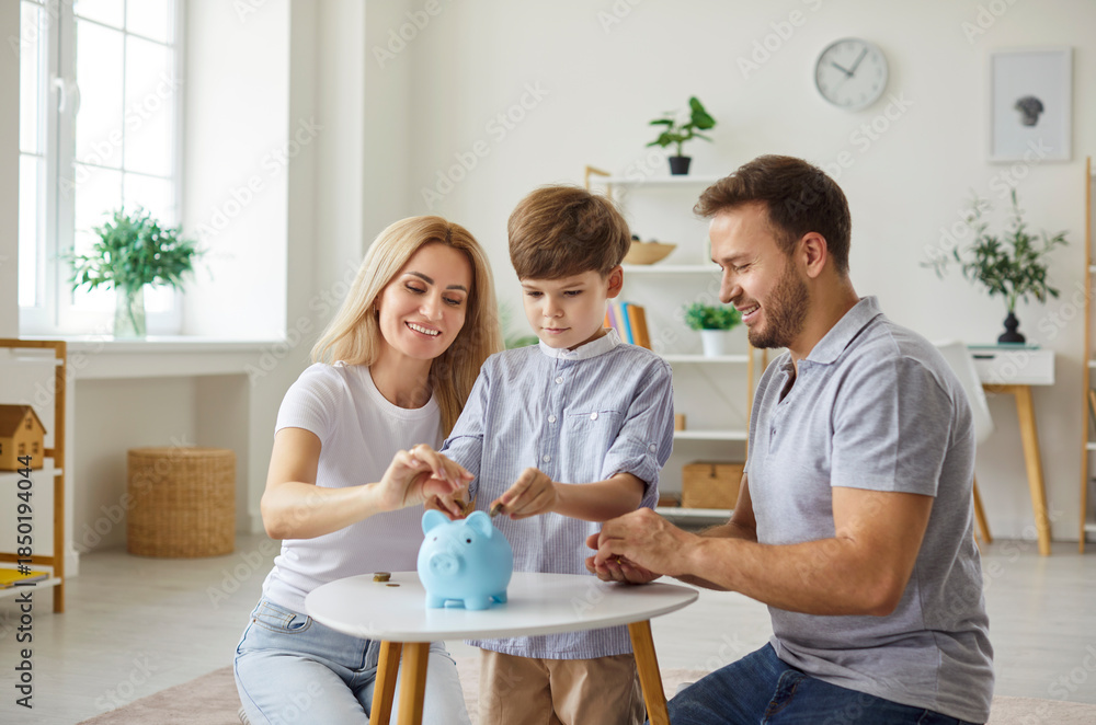 Fototapeta premium Happy young family parents and child sitting on floor in living room and putting coins in blue piggy bank standing on table at home. Smiling mother, father and son planning family budget together.