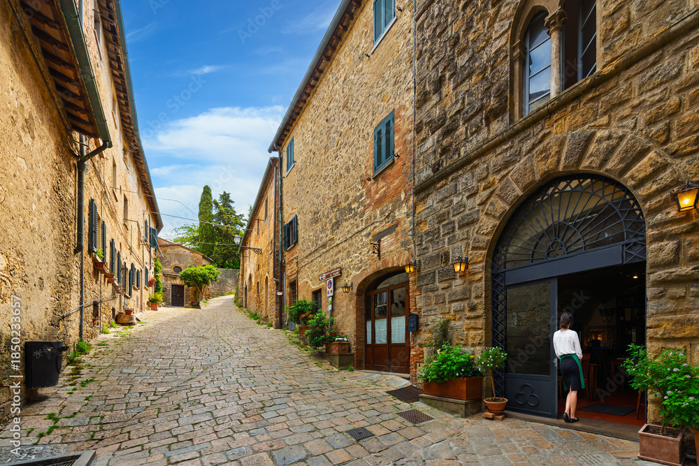 Fototapeta premium A young female server waits in a doorway on a picturesque cobblestone alley in the medieval hill town of Volterra, Italy, Tuscany region.