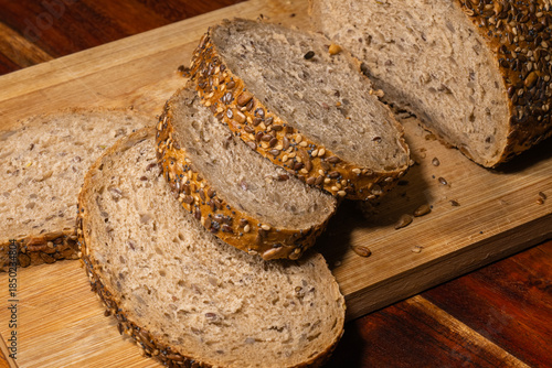 A close-up view of a loaf of seeded whole grain bread with several slices cut, resting on a wooden cutting board with scattered crumbs.