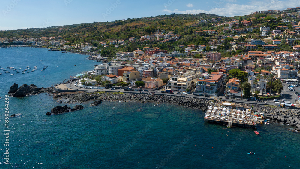 Obraz premium Aerial view of the Aci Trezza waterfront overlooking the Mediterranean Sea, in the province of Catania, Sicily, Italy. In the background is the center of the town. It's a sunny summer morning.