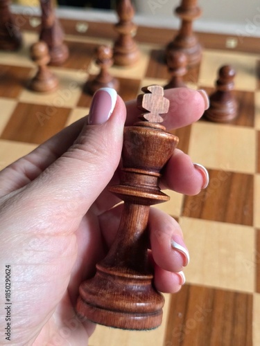 A hand holding a wooden chess piece (king), with a classic French manicure on the nails. Blurred image, business woman hand holding chess price on chess board. Competition success and strategy game