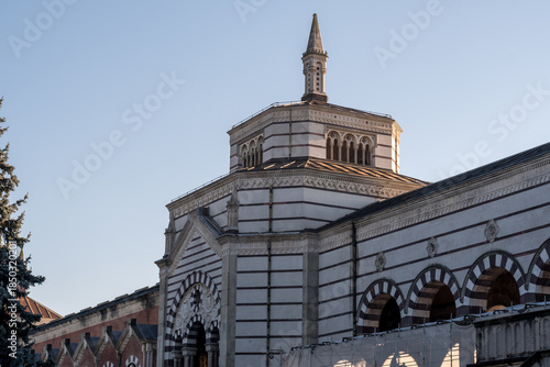 Side view of Famedio chapel dome and striped marble arcades at Monumental Cemetery in Milan, Italy, under clear evening sky.