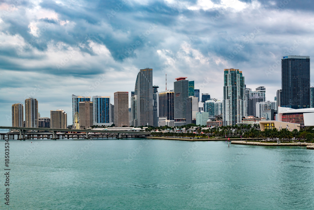 Fototapeta premium The modern Downtown Miami skyline rises dramatically above the turquoise waters of Biscayne Bay on a cloudy day.